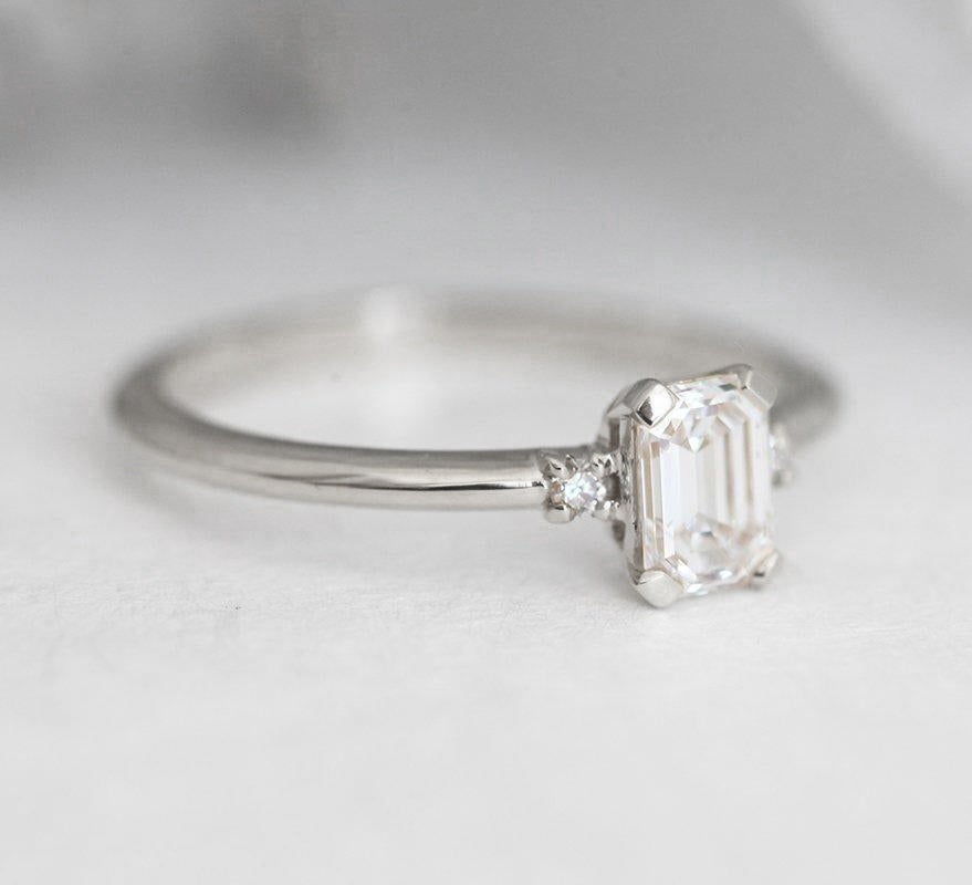 Smaragdno brušeni dijamantski prsten sa bočnim dijamantima na beloj površini, pogled izbliza. Elegantan dizajn sa 3 kamena od 14k žutog zlata.